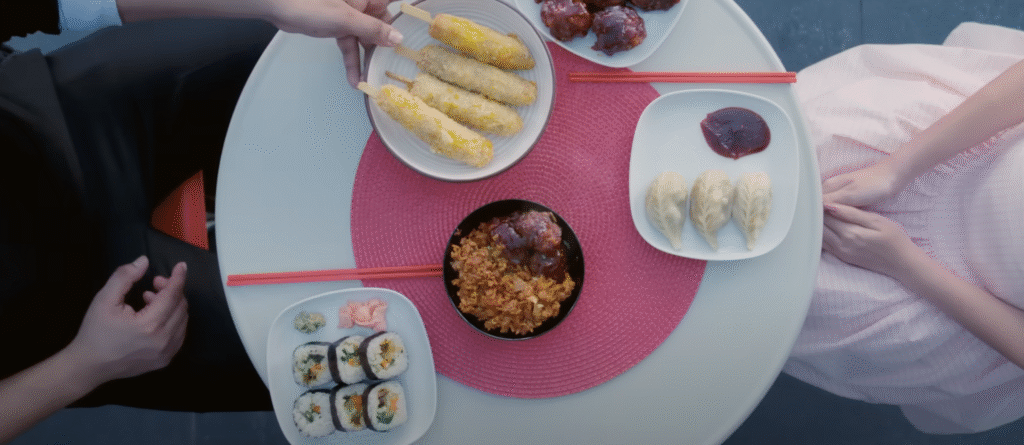 Top-down view of a table set with a variety of Asian dishes including kimbap, steamed dumplings with sauce, fried rice with meatballs, Korean corn dogs, and red chopsticks, with two people seated on either. 
